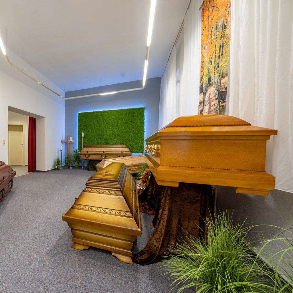 Mehrere helle Särge aus Holz stehen in einem hellen Ausstellungsraum mit grüner Wand und Blumen