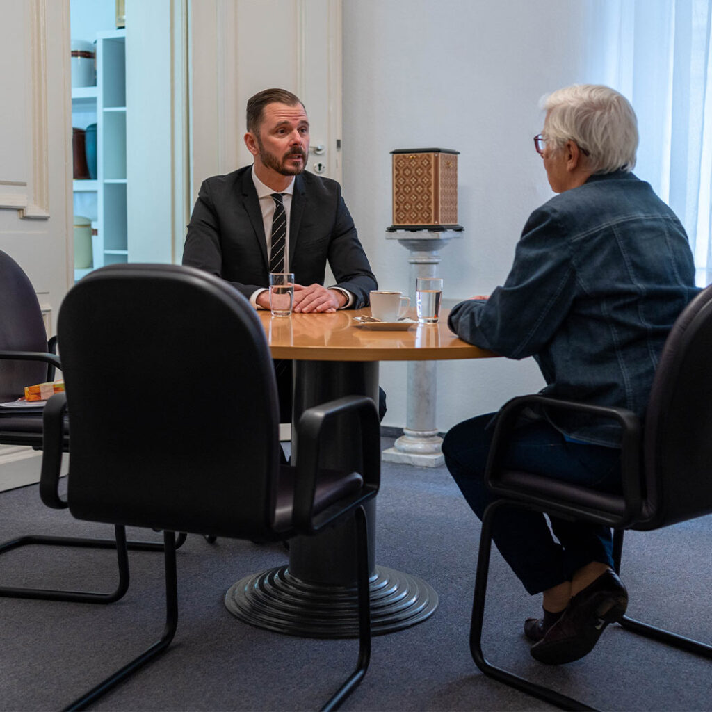 Ein Mann im Anzug spricht ernst mit einer älteren Frau am runden Holztisch in einem Büro