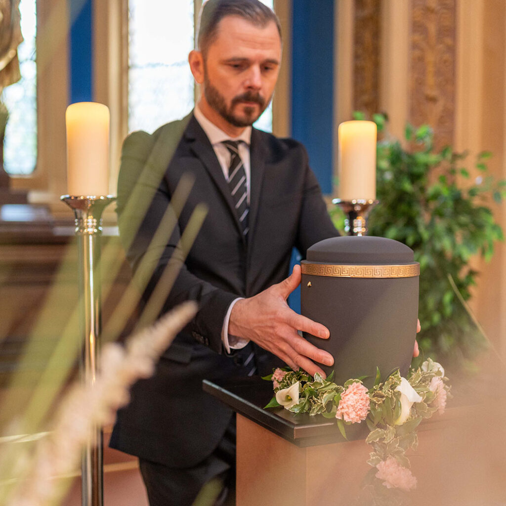 Ein Mann mit Bart und ernster Miene hält eine schwarze Urne mit goldener Verzierung
