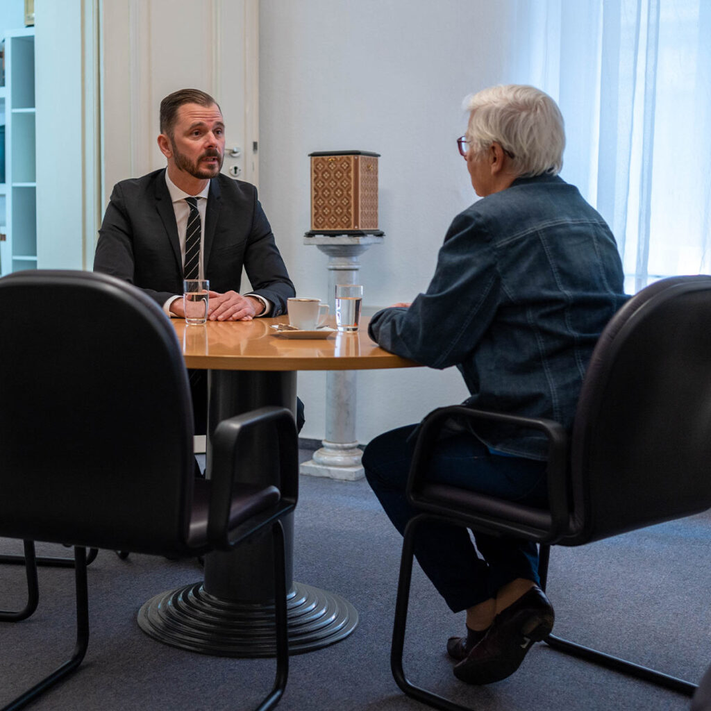 Ein Mann im Anzug spricht ernsthaft mit einer älteren Dame am runden Holztisch