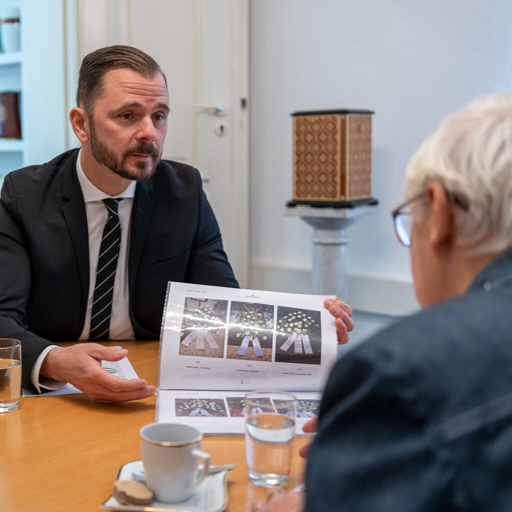 Ein Mann im Anzug zeigt einer älteren Person am Tisch eine bebilderte Broschüre