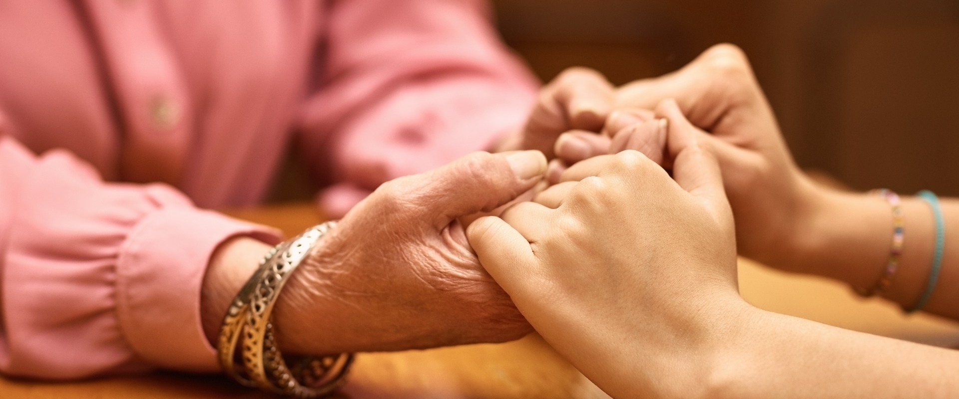 Personen, die sich in einem Pflegeheim an den Händen halten und sich gegenseitig unterstützen, um Hilfe, Empathie oder Hoffnung im Wohnzimmer zu finden. Eine Person, eine Frau oder eine Seniorin an einem Holztisch, die Trauerbegleitung, Vertrauen oder psychologische Betreuung im Ruhestand erhält.
