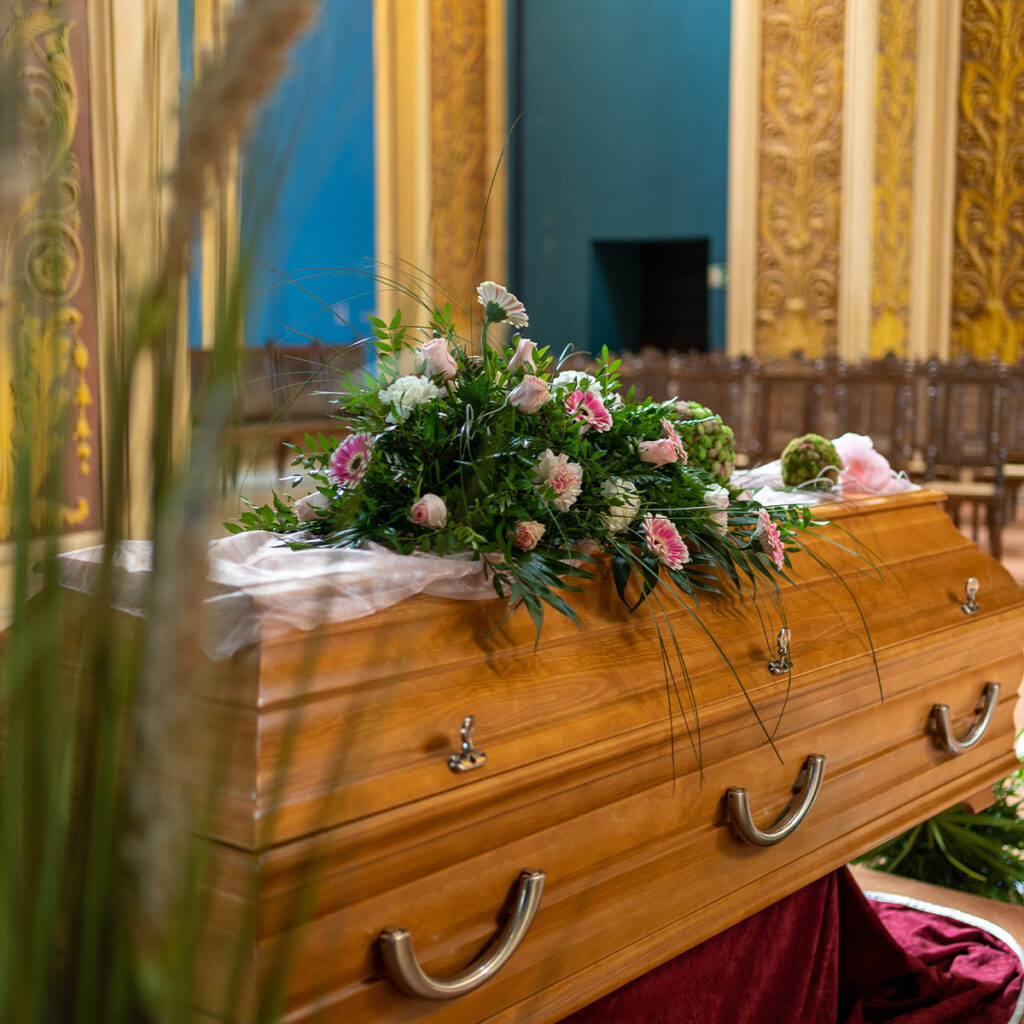 Ein geschlossener Sarg mit Blumengesteck steht in einem prunkvollen Raum