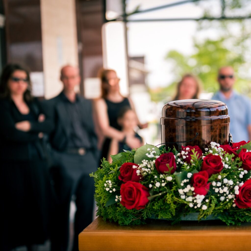 Urne mit Blumenschmuck im Vordergrund, dahinter die Trauergäste